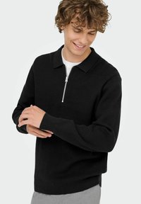 Schwarzer, gerippter Strickpullover mit Reißverschlusskragen, langen Ärmeln und einer lockeren Passform, kombiniert mit einem weißen T-Shirt darunter.