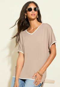 Beige V-hals top met witte afwerking, korte mouwen en een ontspannen pasvorm. Gecombineerd met lichtblauwe denim jeans en gouden accessoires.