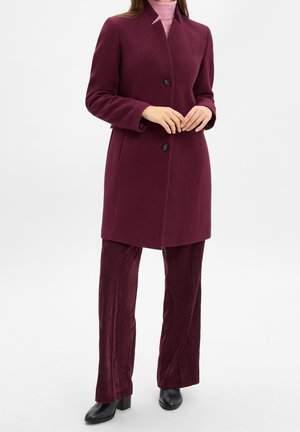Femme portant un manteau bordeaux sur un col roulé rose clair, avec un pantalon en velours côtelé bordeaux assorti et des chaussures à talons noirs.