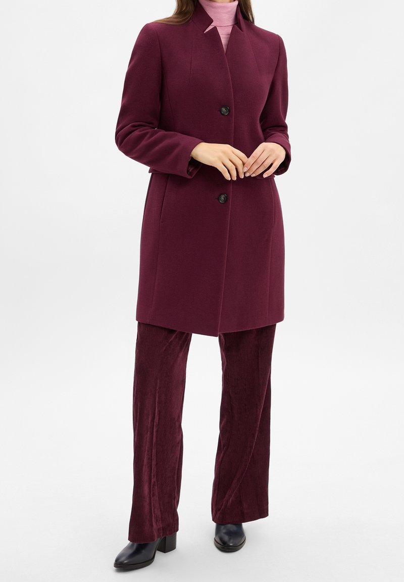 Femme portant un manteau bordeaux sur un col roulé rose clair, avec un pantalon en velours côtelé bordeaux assorti et des chaussures à talons noirs.