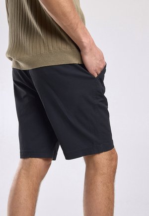 Man draagt zwarte knielange shorts en een beige geribd mouwloos topje, met rechterhand in de zak, staand tegen een effen achtergrond.