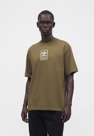 Άνδρας φοράει ελιά πράσινη μπλούζα Adidas με λευκή ετικέτα λογοτύπου στο στήθος και μαύρο παντελόνι, στέκεται μπροστά σε απλό λευκό φόντο.