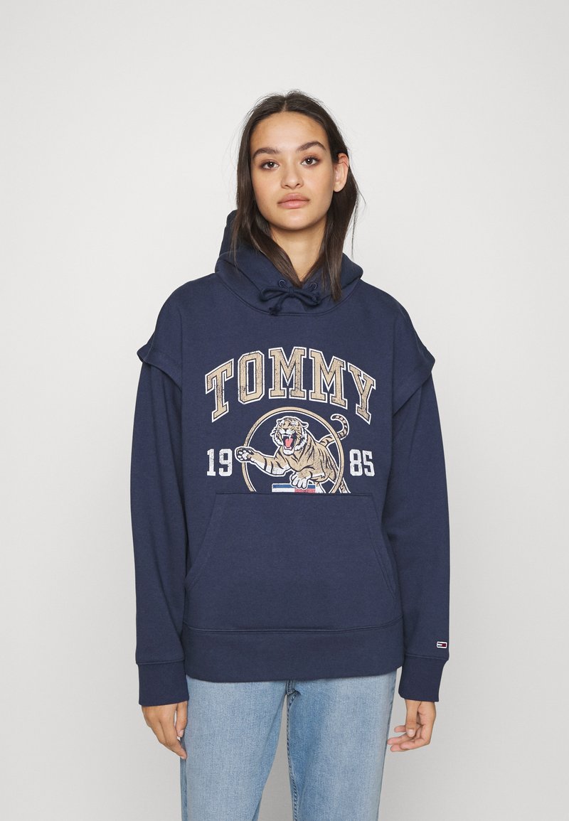 Felpa blu navy con tasca a marsupio, caratterizzata da un grande logo "TOMMY" e una grafica di una tigre. Tessuto morbido, vestibilità comoda e cappuccio regolabile con cordino.
