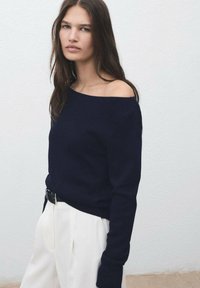 Femme aux longs cheveux bruns portant un pull navy à épaules dénudées et un pantalon blanc large devant un mur blanc uni.