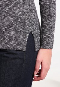 Grauer und schwarzer gestrickter Pullover mit einem strukturierten Muster und seitlichen Schlitzen; über dunklem Denim getragen. Hand an der Seite sichtbar.