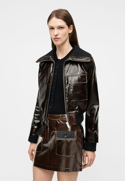 Femme portant une veste zippée brun foncé brillante et une jupe assortie avec de grandes poches avant, par-dessus une chemise noire avec des boutons.