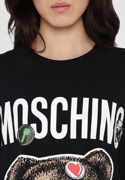 Μαύρο βαμβακερό μπλουζάκι με γραφικό αρκούδας, μεγάλο λογότυπο "MOSCHINO" και κυκλικά πατρόν με καρδιά και μπρόκολο. Κλασική σχεδίαση με στρογγυλή λαιμόκοψη.