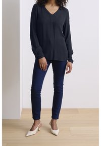 Blouse noire à manches longues et col en V en tissu léger ; associée à un jean bleu foncé ajusté et des escarpins slingback ivoire à bouts pointus.