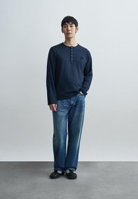 Μπλε ναυτικό μακρυμάνικο πουκάμισο henley με πατιλέτα κουμπιών, φορεμένο με ανοιχτό μπλε τζιν παντελόνι με φαρδιά μπατζάκια και μαύρα αθλητικά παπούτσια με λευκές ρίγες.