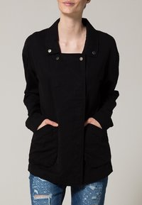 Giacca nera in cotone con scollatura quadrata, chiusure a scatto e ampie tasche frontali. Abbinata a jeans blu strappati.