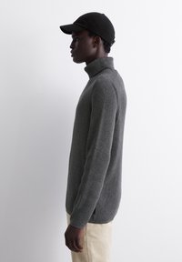 Grauer gerippter Rollkragenpullover aus strukturiertem Stoff, mit langen Ärmeln und seitlichen Schlitzen. Kombiniert mit einer schwarzen Mütze und hellen Hosen.