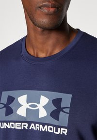 Marineblaue Baumwoll-Sport-T-Shirt mit einem grauen rechteckigen Grafik, die drei große Logos zeigt, eines in Weiß und zwei in Dunkelblau. Crew-Neck-Design.