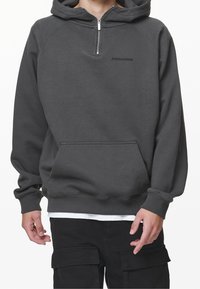 Man bär en mörkgrå huppartröja med dragkedja fram och ficka, kombinerad med svarta cargobyxor och en vit undertröja som syns nedtill.