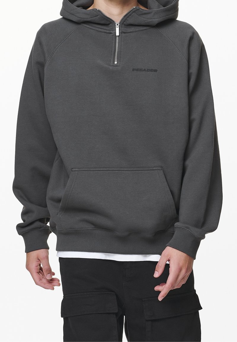 Man bär en mörkgrå huppartröja med dragkedja fram och ficka, kombinerad med svarta cargobyxor och en vit undertröja som syns nedtill.