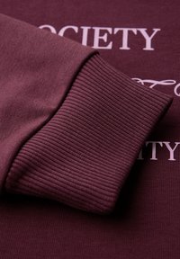 Sweatshirt bordeaux avec des poignets côtelés et un tissu lisse. Texte "SOCIÉTÉ" en rose clair sur la poitrine. Vue rapprochée des détails.