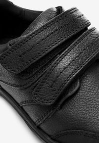 Zwarte leren schoen met twee verstelbare klittenbandsluitingen, gestructureerd oppervlak, versterkte stiksels en een rond neusontwerp.