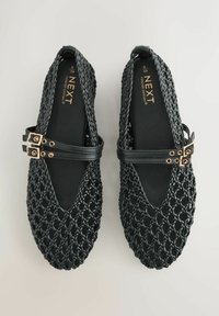 Schwarze gewebte Schuhe mit einem strukturierten Design und einer V-förmigen Zehenpartie, gesichert mit zwei Riemen mit goldfarbener Schnalle. Glattes Innenfutter.