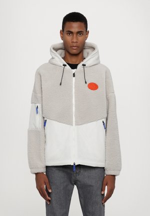 Giacca in pile grigia con la parte inferiore bianca, cappuccio attaccato, zip blu e un logo circolare arancione. Presenta una finitura testurizzata e un fit rilassato.
