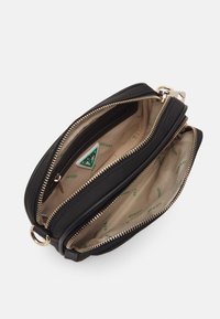 Svart läder crossbody-väska med guld blixtlås och en beige tyginteriör, med en triangulär Guess-logotag och flera fack.