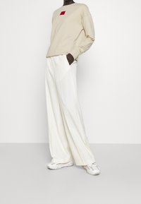 Beżowa bluza z czerwoną kwadratową grafiką, zestawiona z białymi spodniami o szerokich nogawkach. Model ma na sobie białe trampki w wzory. Gładkie, casualowe materiały.