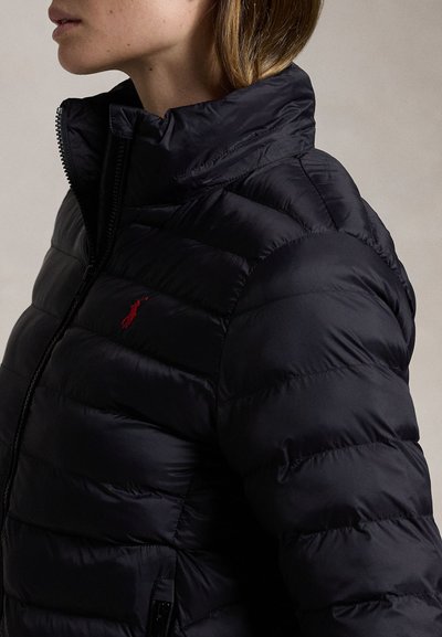 Žena nosící černou vyteplenou bundu s červeným vyšívaným logem na hrudi, stojící z profilu na neutrálním pozadí.