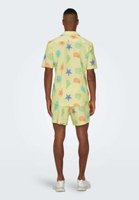 Camisa de manga corta amarilla y pantalones cortos a juego, con patrones coloridos de mar, como conchas y estrellas. La tela parece ligera y texturada.