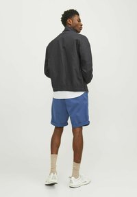 Veste bomber légère noire avec un col haut, associée à un short bleu retroussé. Chemise blanche visible en dessous, chaussettes de couleur neutre et baskets.