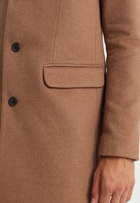 Cappotto in misto lana marrone con revers a punte, grandi bottoni e una tasca frontale. Il tessuto ha una consistenza liscia e una finitura morbida.
