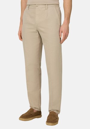 Homme portant un pantalon beige à jambes droites et des chaussures marron en daim à enfiler, debout devant un fond uni.