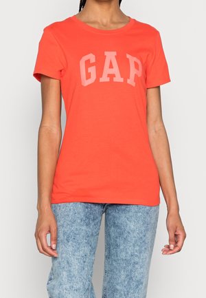 Person iført en klar orange kortærmet trøje med "GAP"-logo og lyseblå acid wash-jeans, stående mod en ensfarvet baggrund.