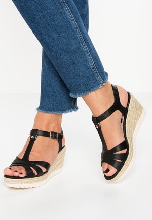 Fødder iført sorte læder kilehælssandaler med ankelremme og blå jeans med frynset kant på hvid baggrund.