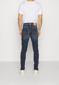 Jeans en denim bleu foncé avec une coupe slim, dotés de poches arrière appliquées et d'une étiquette de marque sur la taille. Portés avec un t-shirt blanc uni.