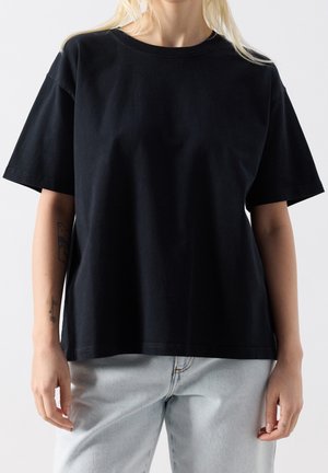 Vaaleahiuksinen nainen, joka käyttää yksinkertaista mustaa ylip-sized t-paitaa ja vaaleansinisiä farkkuja, seisoo käsivarret rentoina sivuilla.