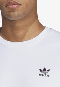 Bílá bavlněná mikina s kulatým výstřihem a žebrovanými detaily. Na levé straně hrudníku je malý černý logo Adidas.