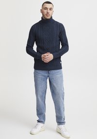 Un pull à col roulé en maille bleu marine avec un motif de cable texturé, des poignets et un ourlet côtelés. Associé à un jean en denim bleu clair et des chaussures blanches.