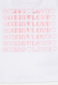 Weißes Baumwoll-T-Shirt mit mehrfach wiederholtem "GUESS ♥ LOVE"-Text in verschiedenen Rosatönen, wobei einige Buchstaben texturiert sind, um einen Kontrast zu erzeugen.