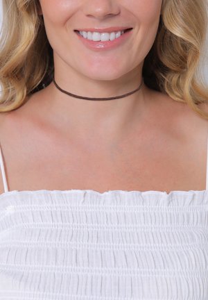 Brun choker halskæde lavet af læder, med et simpelt, tyndt design der hviler tæt på halsen. Båret med en hvid tekstureret top.