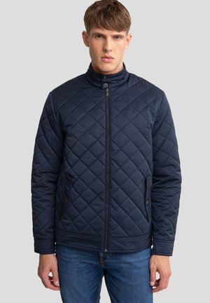 Navy quilted jacket with a stand collar, front zip, and two side pockets. Features diamond pattern and textured fabric. Worn with blue jeans.