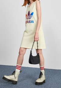 Żółta sukienka bez rękawów z dużym, wielokolorowym logo Adidas, zestawiona z białymi botkami, skarpetkami z czerwonymi akcentami i małą czarną torbą.