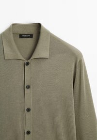 Chemise tricotée vert olive à boutons avec col, comportant quatre boutons noirs sur le devant, posée à plat sur un fond blanc.