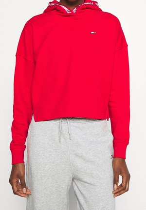Persoon draagt een rode cropped hoodie met logo op de borst en een gebrande capuchon, gecombineerd met lichtgrijze sweatpants met trekkoord.
