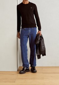 Czarny sweter z długim rękawem z małym logo, niebieskie dżinsy o prostych nogawkach oraz czarne skórzane buty. Model trzyma czarną skórzaną kurtkę.