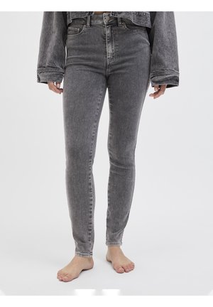 Osoba ubrana w wysokie, wyblakłe czarne obcisłe dżinsy i dopasowaną kurtkę jeansową, stojąca boso na jednolitym białym tle.