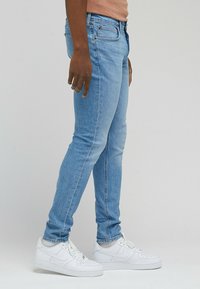 Jeans in denim azzurro chiaro con una vestibilità slim, cinque tasche e dettagli di cucitura discreti, abbinati a sneakers bianche.