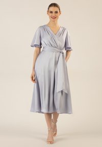 Ein fliederfarbendes Satin-Wickelkleid mit V-Ausschnitt, kurzen flatternden Ärmeln und einem gebundenen Taillenbereich. Es hat einen fließenden Rock und Taschen. Dazu silberne Riemchen-High Heels.