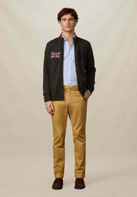 Mladý muž v černé zipové mikině s nášivkou Union Jack, světle modré košili, hořčicových kalhotách a tmavě hnědých botách, stojící proti béžové zdi.