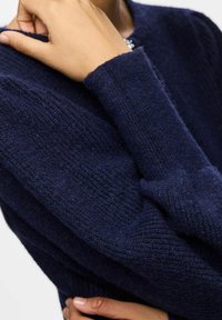 Jūras zila adīta džemperis ar strukturētu rakstu, apaļu apkakli un garām piedurknēm. Materiāls izskatās mīksts un nedaudz pūkains. Roka novietota uz pleca.