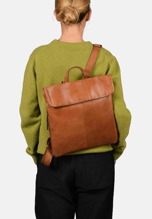 Sac à dos en cuir marron avec un rabat, de forme rectangulaire, et une poignée supérieure. La sangle est réglable, présentée sur un fond de pull vert.