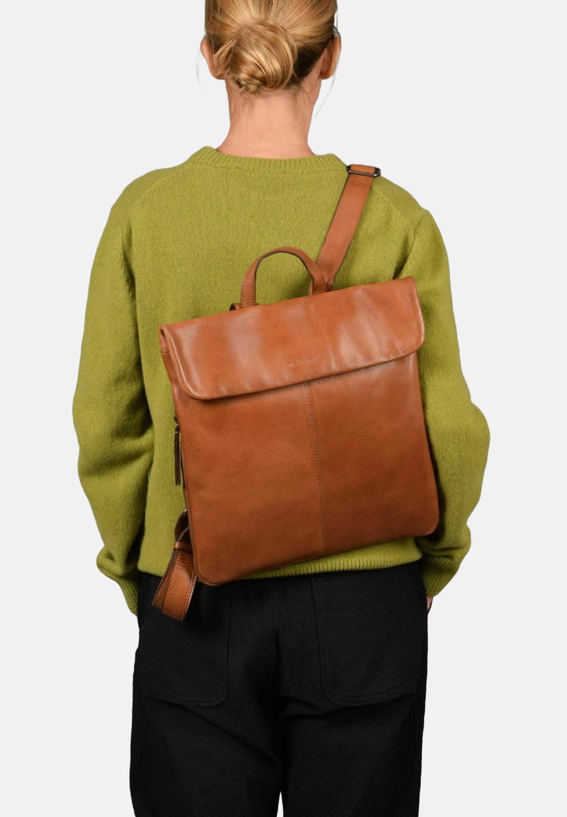 Mochila de cuero marrón con tapa, de forma rectangular y con un asa en la parte superior. La correa es ajustable, colocada sobre un fondo de suéter verde.
