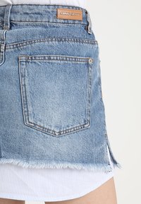 Gonna mini in denim azzurro chiaro con orlo sfilacciato, design a cinque tasche e etichetta in pelle marrone sul retro, con dettagli di sfumature sottili.